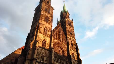 slow pan and tilt of the historic chruch lorenzkirche in nuremberg. the two high towers are shining golden in the last sunlight of the day at sunset while many people are busy walking in the pedestrian zone below.