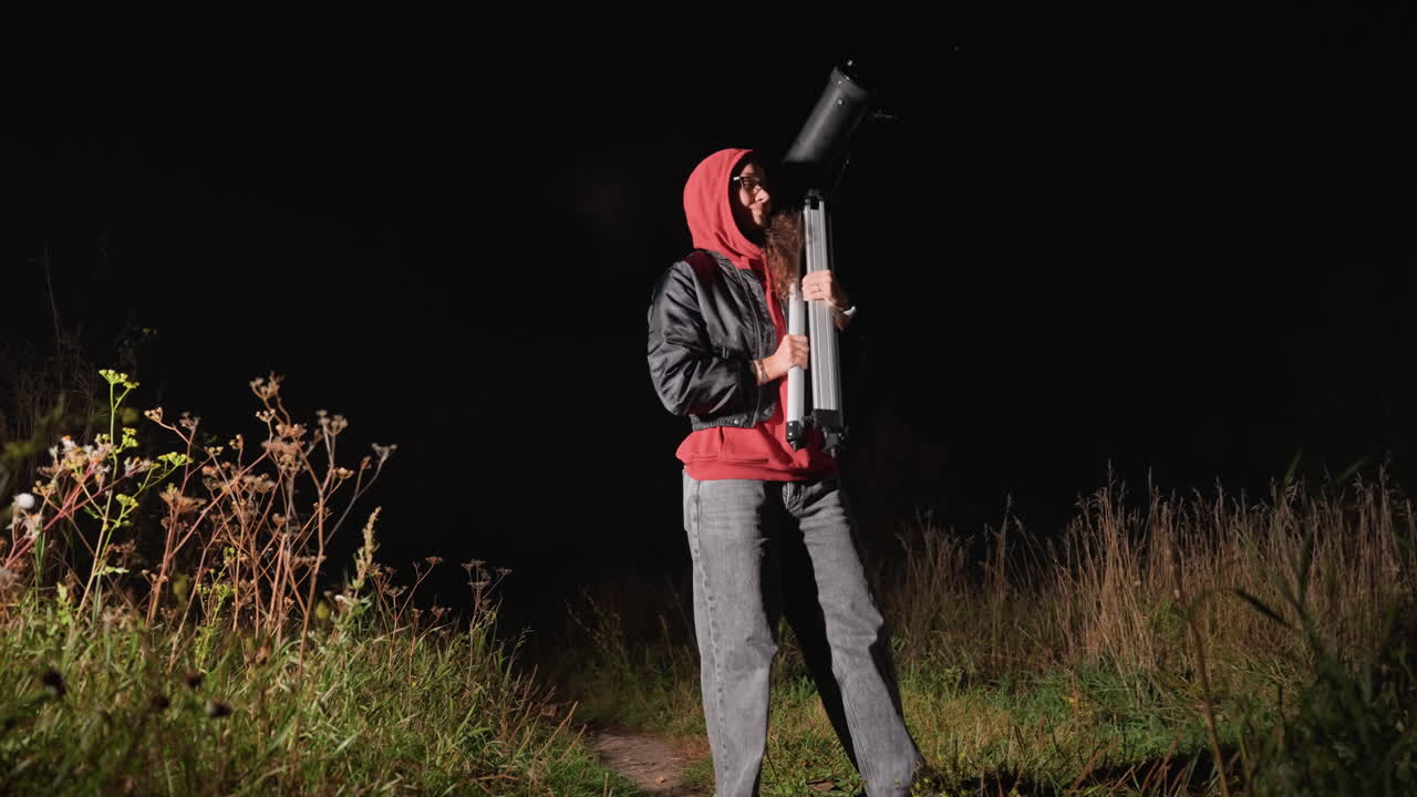 Night sky stargazing with telescope, outdoors person in red hoodie adjusts tripodmounted refractor on grassy meadow path, focused on distant stars, quiet atmosphere with soft flashlight glow