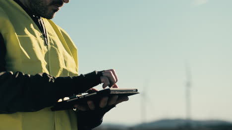 un primer plano de un joven ingeniero de energía renovable que usa una tableta para comprobar las turbinas eólicas en un campo, promoviendo el uso de fuentes de energía sostenibles