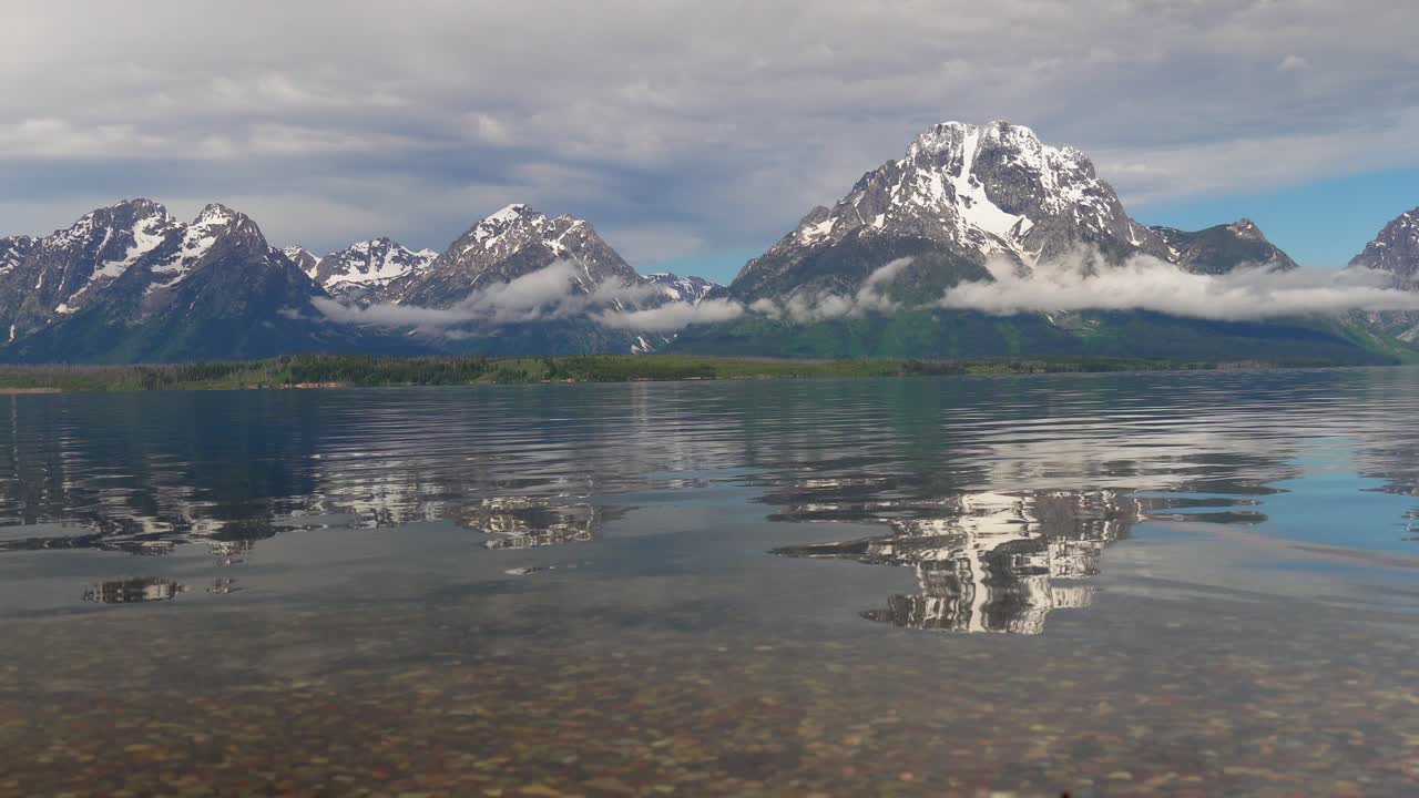 다채로운 자갈의 잭슨 호수 해안과 백그라운드에서 테톤이 있는 그랜드 테톤 국립공원의 졸졸 흐르는 물을 볼 수 있습니다.