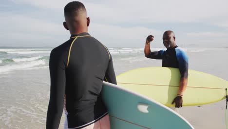 Afroamerikanischer-Vater-Und-Sohn-Im-Teenageralter-Stehen-Am-Strand,-Halten-Surfbretter-In-Der-Hand-Und-Reden