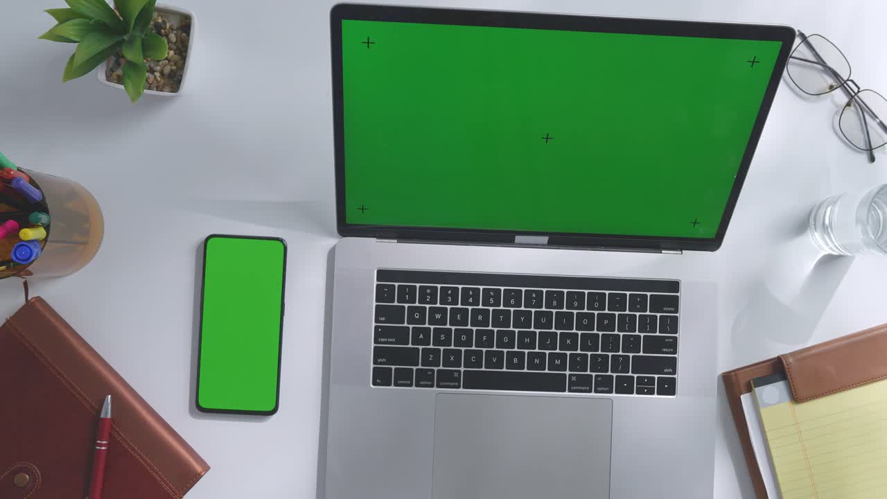 vista de arriba hacia abajo de una computadora portátil con pantalla verde simulada pantalla chromakey en un escritorio de oficina de madera al lado de la libreta con bolígrafos, gafas y un vaso de agua.