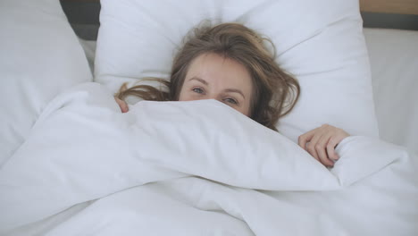 mujer feliz asomándose de su manta en la cama y sonriendo. asomándose debajo del edredón y sonriendo