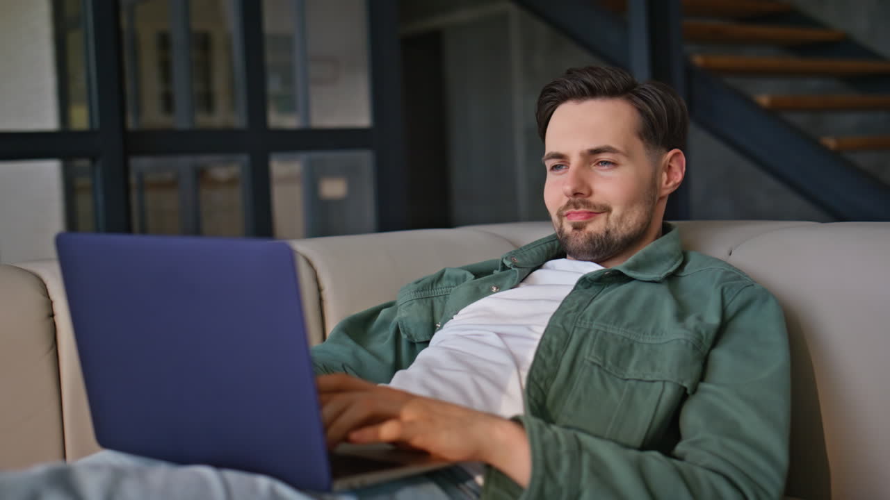 Closeup man working laptop lying at light couch. Relaxed guy typing computer