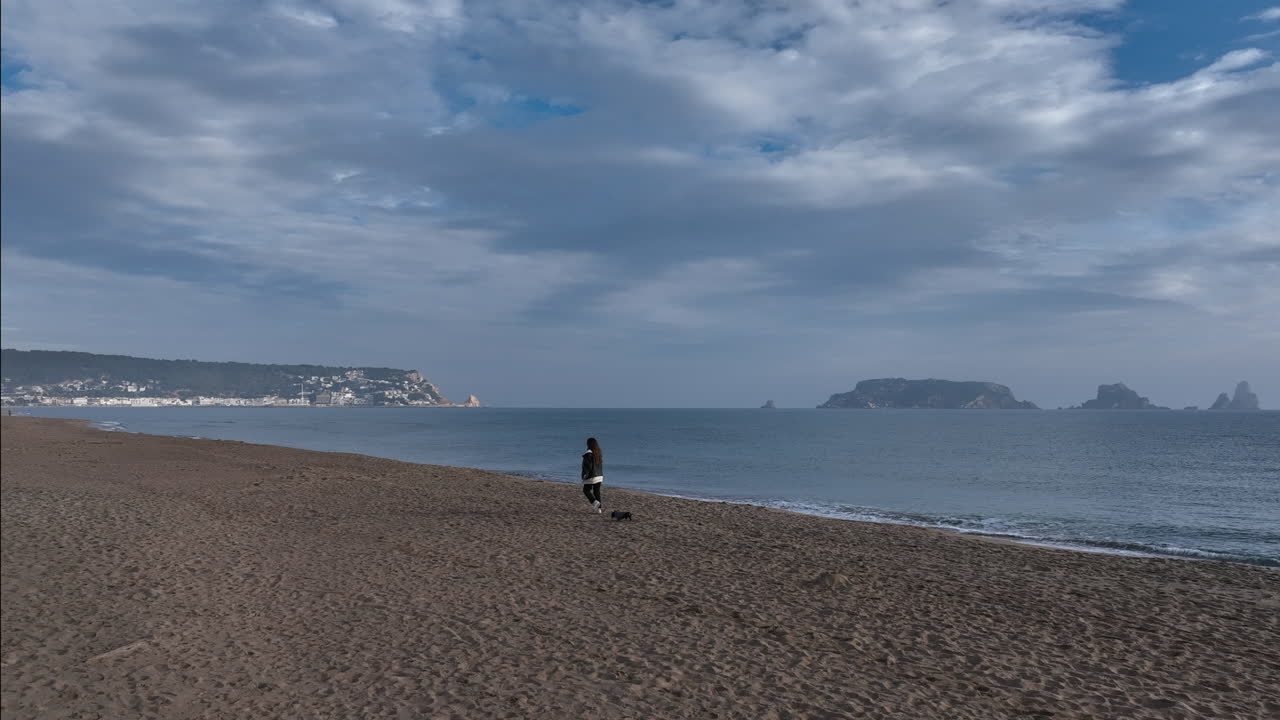 mujer caminando dachshund en miniatura a través de la playa vacía con vistas a la costa de las islas medes al amanecer.