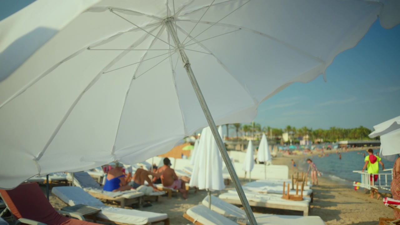 Ombrellone da spiaggia bianco che fornisce ombra su una sdraio, con persone che si rilassano, nuotano e si godono le vacanze estive.