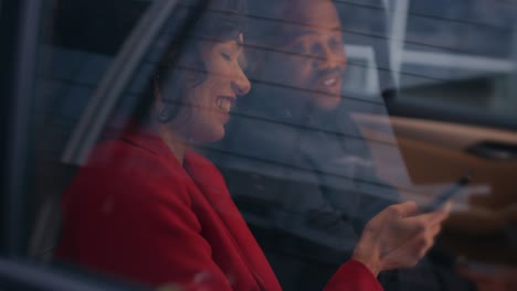 beautiful woman and her male partner riding in the back seat of a car, they share smartphone screen and talk and have fun. it's evening in the big city.