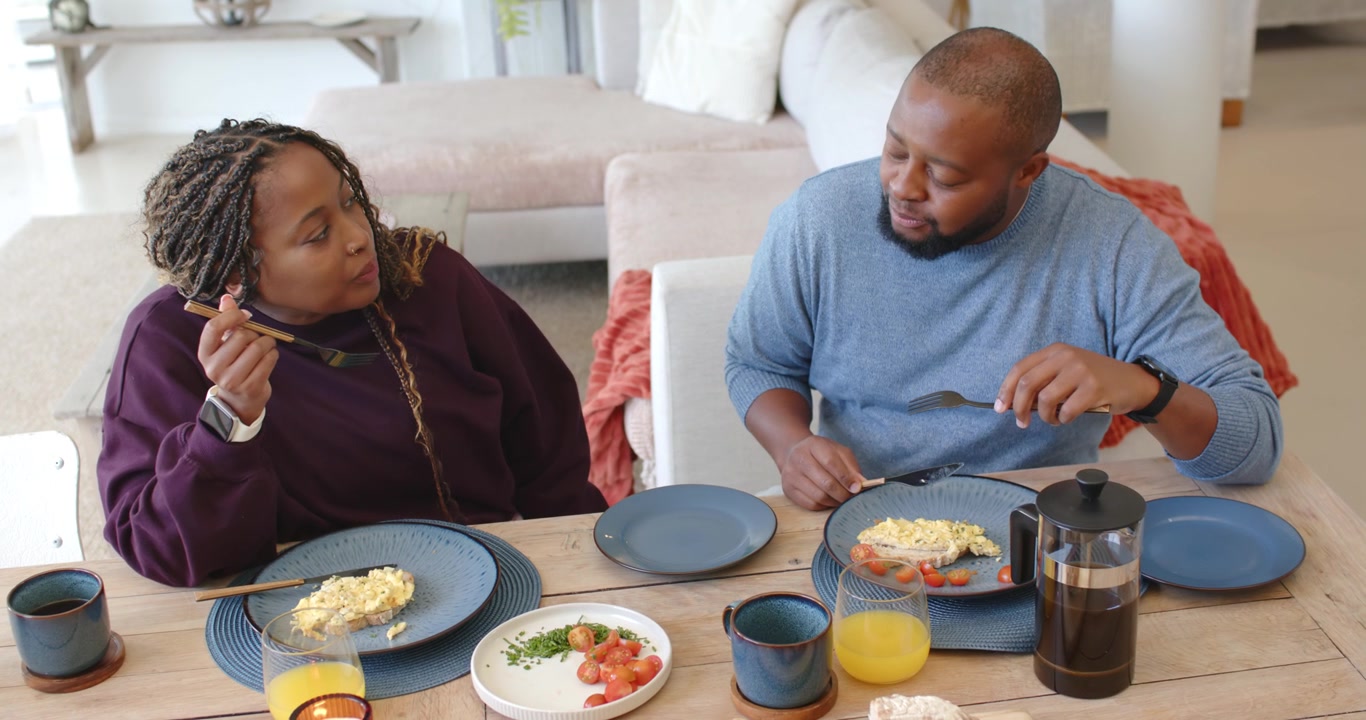 African American couple eating eggs at table while man glancing, gesturing, woman pausing listening