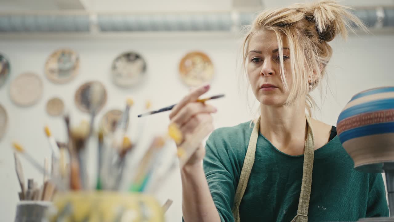 Pottery decoration. Woman artist choosing paintbrush, drawing pattern on handmade ceramic pot at workshop, slow motion