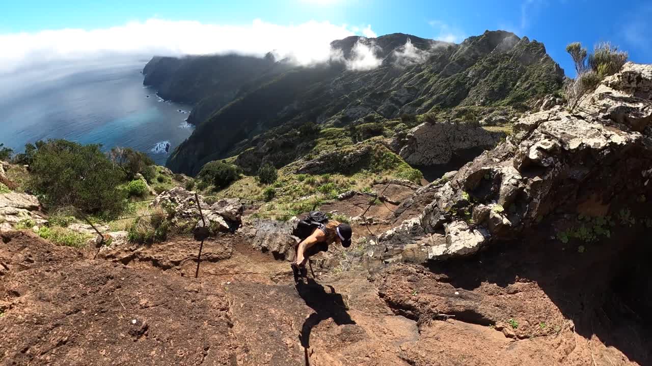 A young and strong solo man hiker is holding a selfie stick with a 360 action camera as he climbs up the rocky cliffs of Espigao Amorelo in Madeira with an amazing and epic view behind him.
