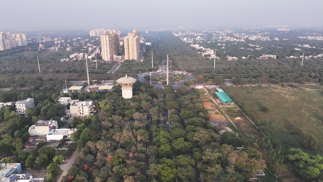 Vista aerea di Greater Noida che mostra una torre dell'acqua che si erge sopra una fitta vegetazione, pali elettrici, grattacieli residenziali in lontananza e una leggera foschia che rivela il paesaggio urbano in espansione della città