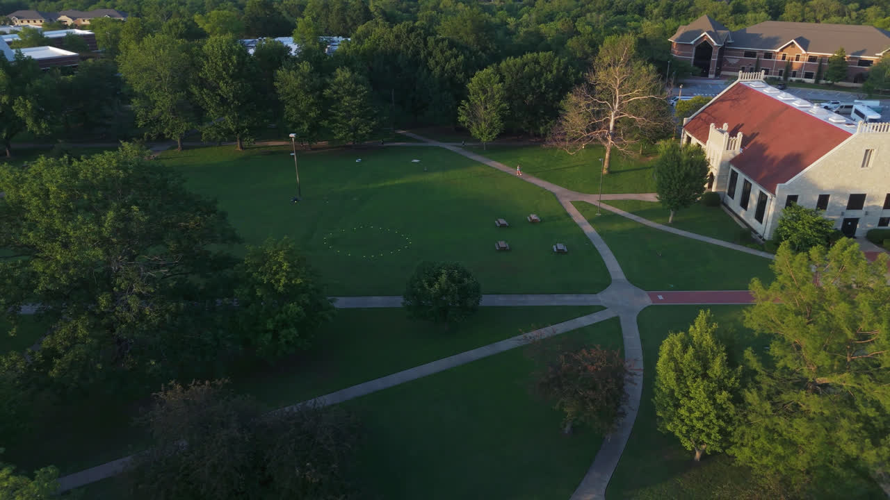 Open campus lawns with nearby academic buildings and tree cover, aerial pullback over John Brown University JBU sign