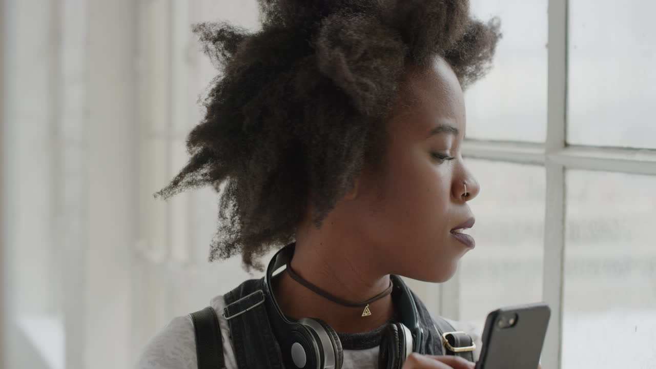 ritratto di una giovane studentessa afroamericana che usa lo smartphone, scrive messaggi, naviga, aspetta vicino alla finestra, annoiata, legge messaggi sms solo sul cellulare