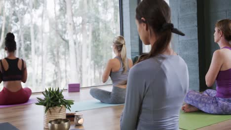 Rear-view-of-diverse-women-practicing-meditation-while-sitting-on-mats-in-yoga-studio