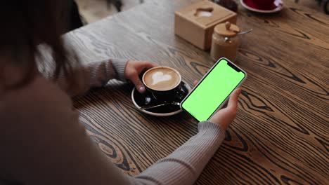 close up back view of female hands holding smartphone with green screen for copy space sitting in coffee shop. chroma key. watching video, surfing internet, social network. modern technology concept