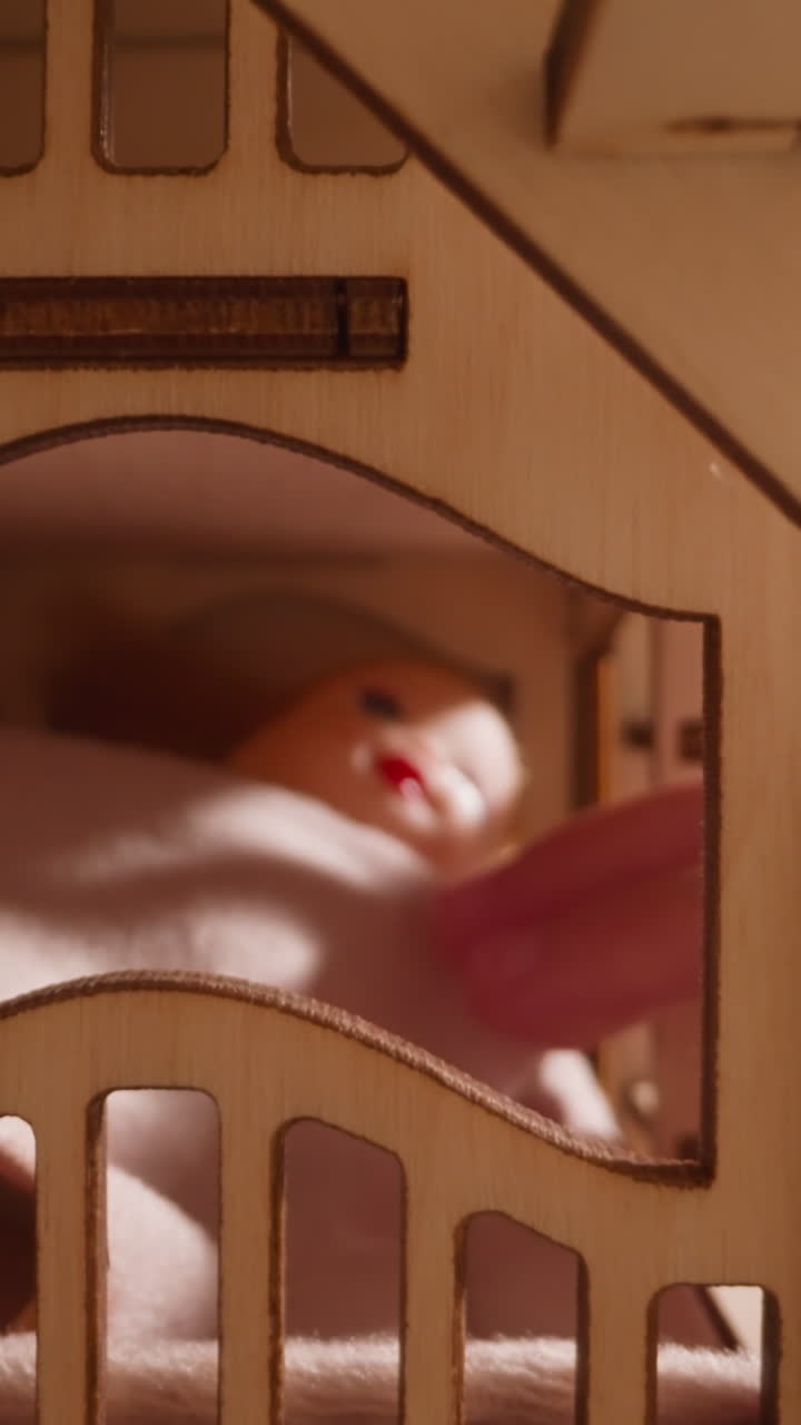 La mano di una bambina tocca e sistema un morbido lenzuolo di stoffa su una bambola sdraiata nel letto in una casa giocattolo, vista ravvicinata con zoom indietro attraverso la stanza con gradini e finestra.
