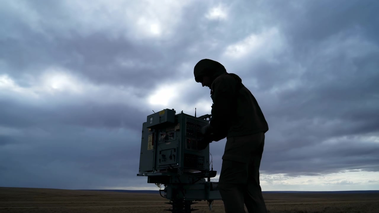 Backlit figure leans toward panel controls and cables on field-mounted equipment while a flat brown landscape stretches to the horizon under dense layered storm clouds.