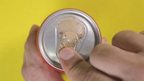 above close-up shot: a hand opening a tin can, unleashing a burst of effervescent gas from a rich dark liquid carbonated drink, then taking the container away