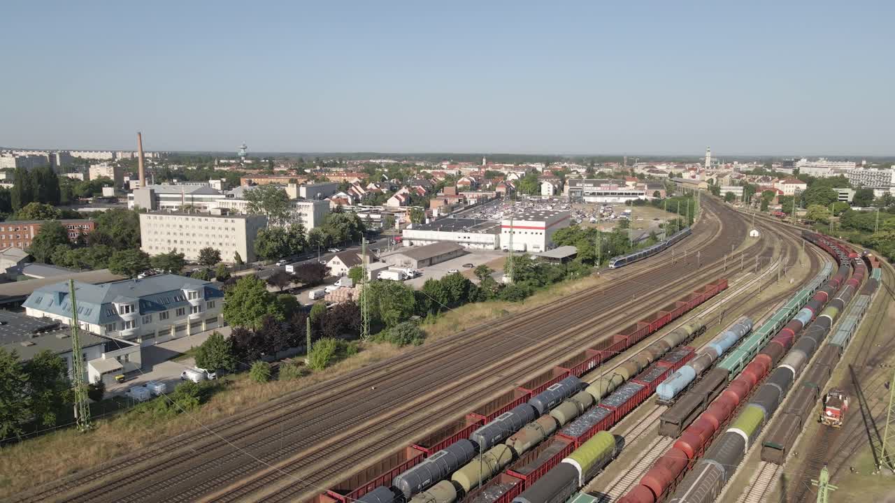 Aerial view of a busy freight train yard filled with various colored cargo wagons. In the background, a vibrant cityscape with residential, industrial, and commercial buildings.