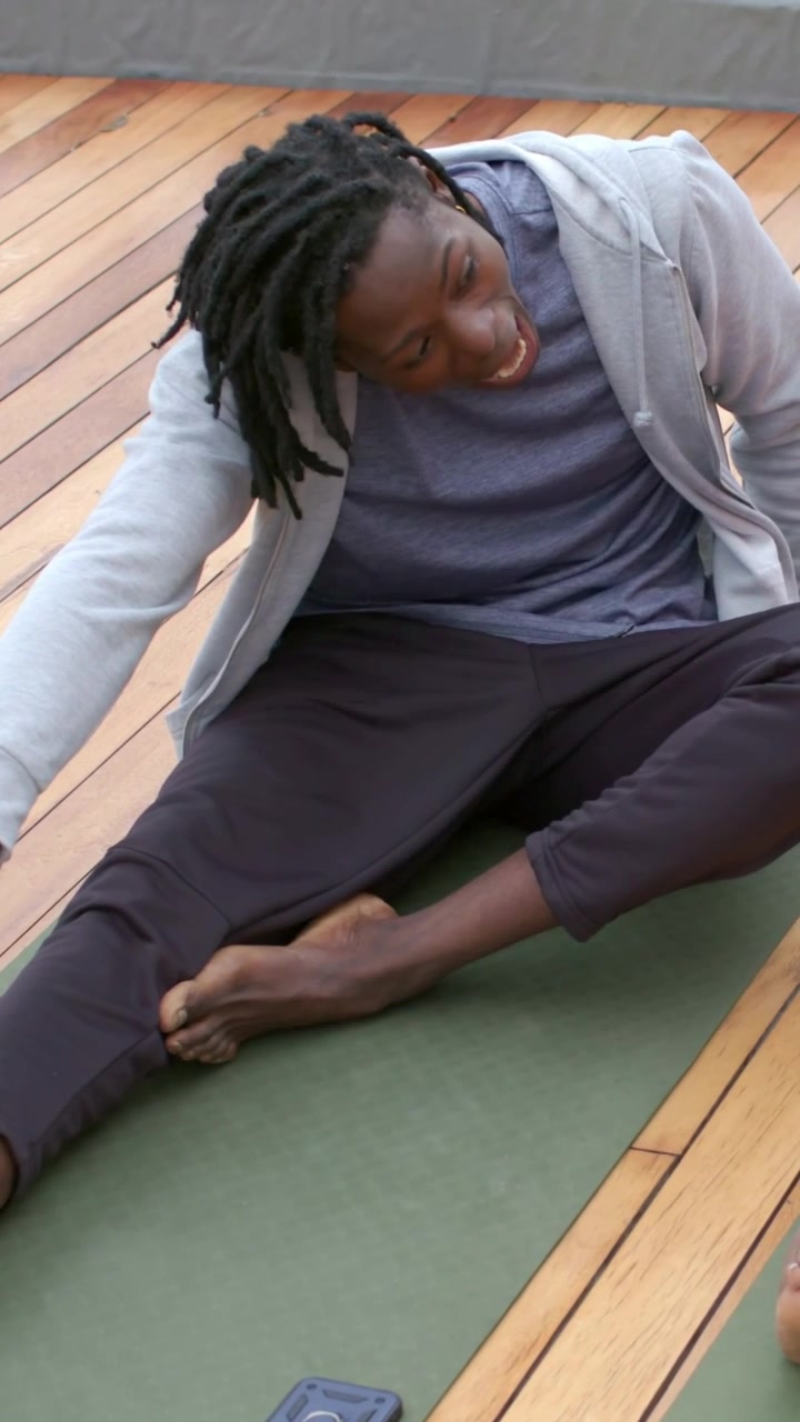 Vertical video: Leaning hoodie man reaching left foot on green mat on deck stretching, phone by mat