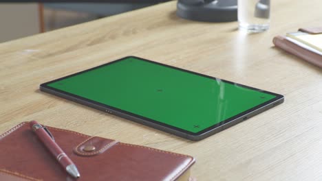 top down view of a laptop computer with mock up green screen chromakey display on a wooden office desk next to notebook with pens, glasses, and a glass of water. slow zoom out, close up