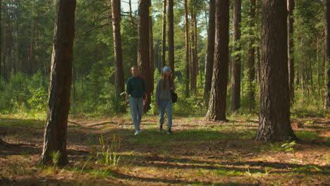 two hikers walking through sunlit forest, one with red backpack and cloth draped over it, other with blue headscarf, gazing thoughtfully to the left, sunlight filters through tall trees