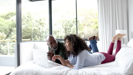 happy mature diverse couple lying on bed using laptop at home, slow motion