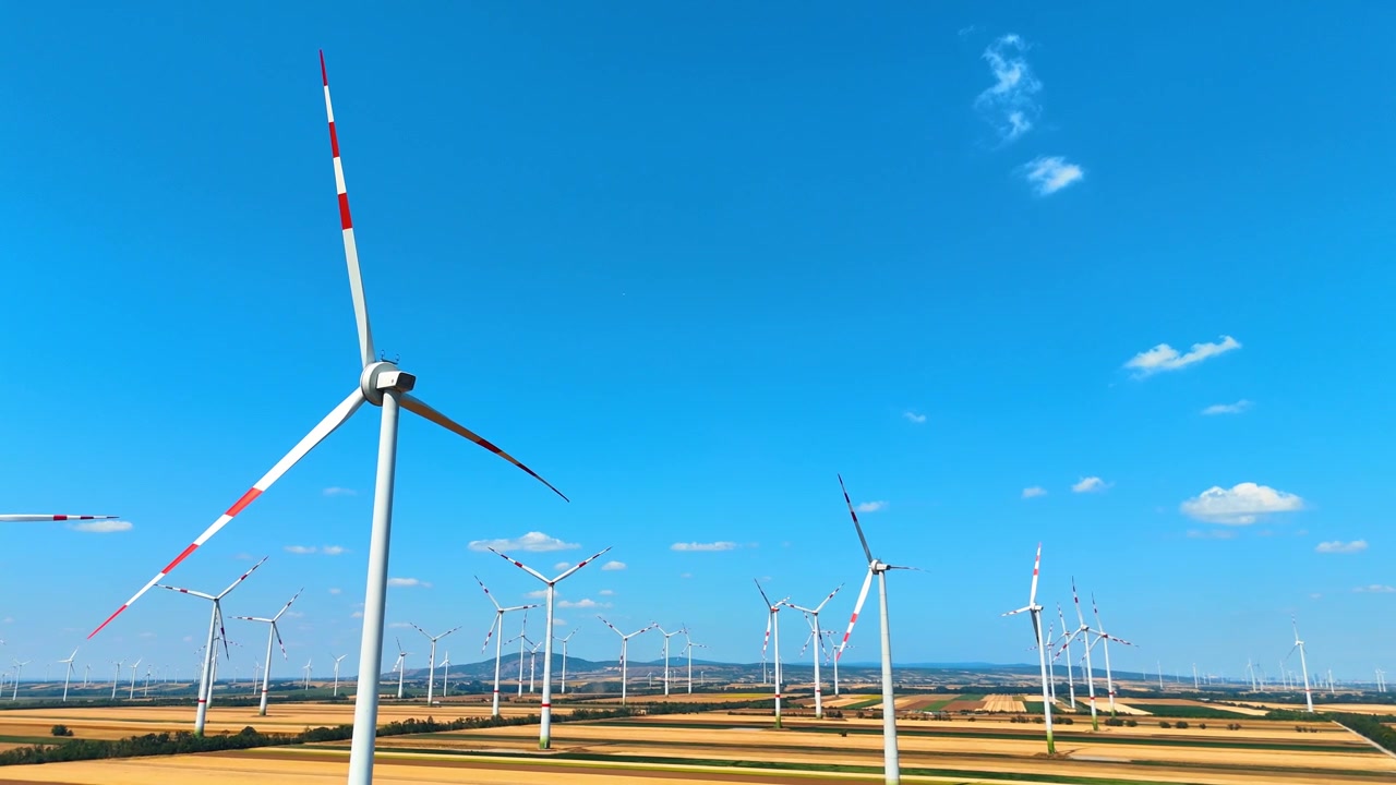 青空の下、黄色い作物が広がる畑に立つ風力発電機。明るい地平線を背景に、回転するブレードを持つ風力発電機のナセルの空撮クローズアップ。