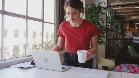 joven hispana entrando a la oficina con una taza y un teléfono y sentada en su escritorio para trabajar, cerca