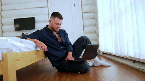 focused man wearing dark shirt sits on the floor leaning on the bed. freelancer looks at the laptop on his knee working remote.