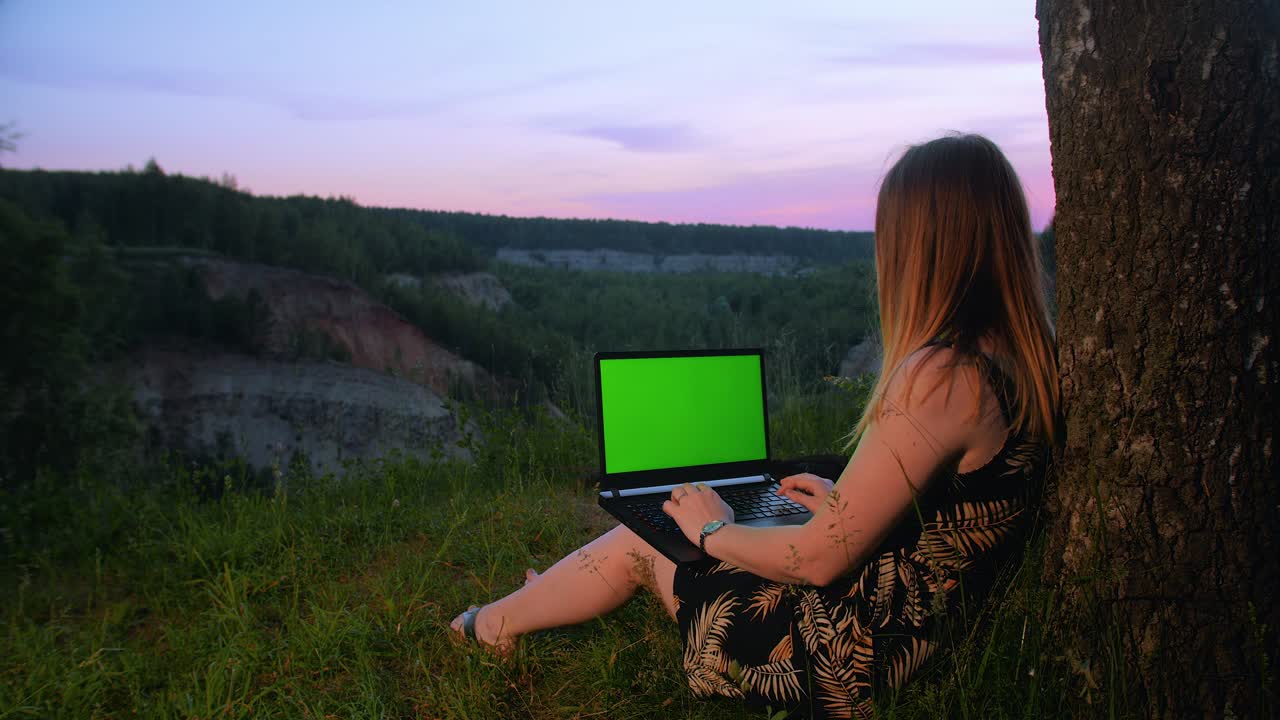 una joven trabajando en una computadora portátil con una pantalla verde cerca de un árbol en la cima de una montaña.