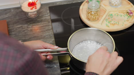 close-up hand view of person stirring sugar in pot on stovetop while beads of different colors are scattered on a wooden tray nearby, kitchen scene focused on cooking sugar syrup