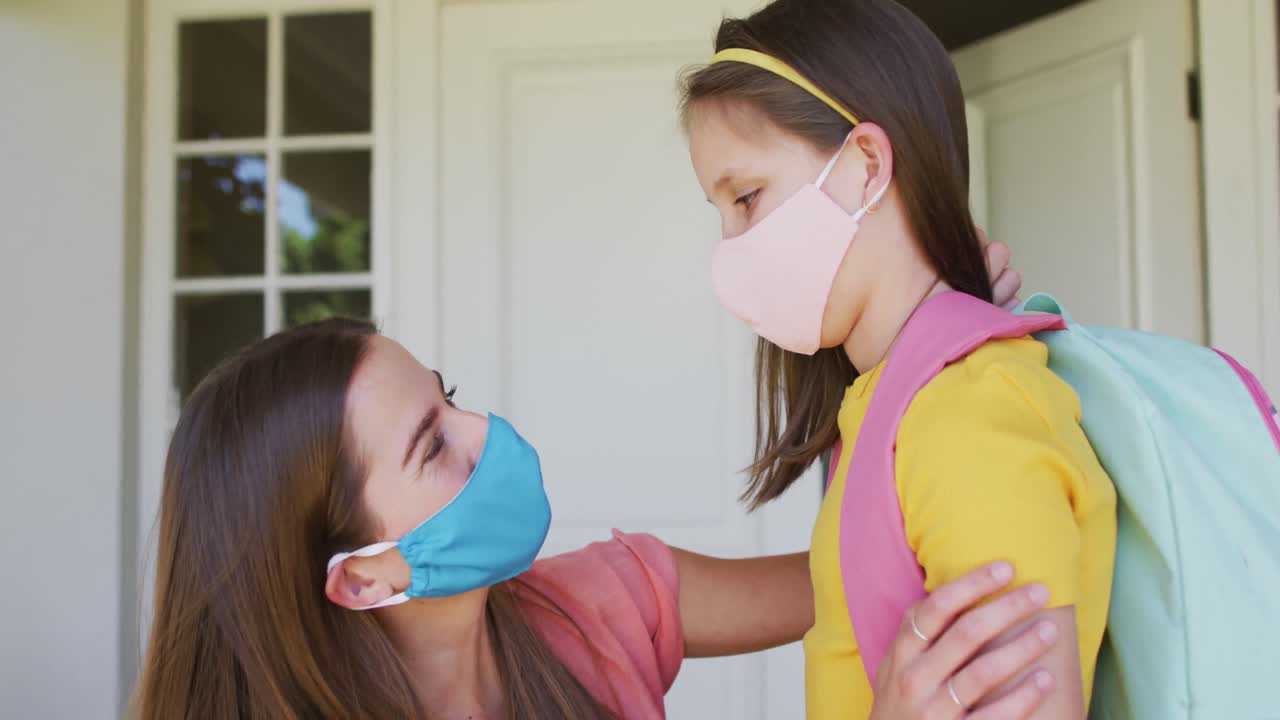 얼굴 마스크를 쓴 백인 어머니가 집 밖에서 딸을 안아주고 있습니다.