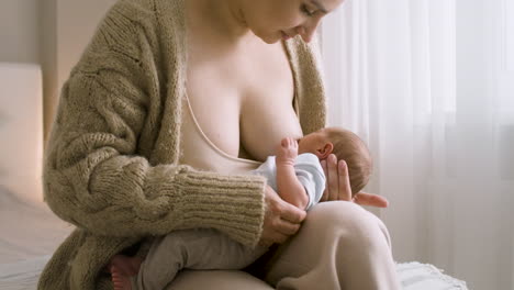 liebevolle junge mutter, die ihr neugeborenes baby stillt, während sie zu hause auf dem bett sitzt