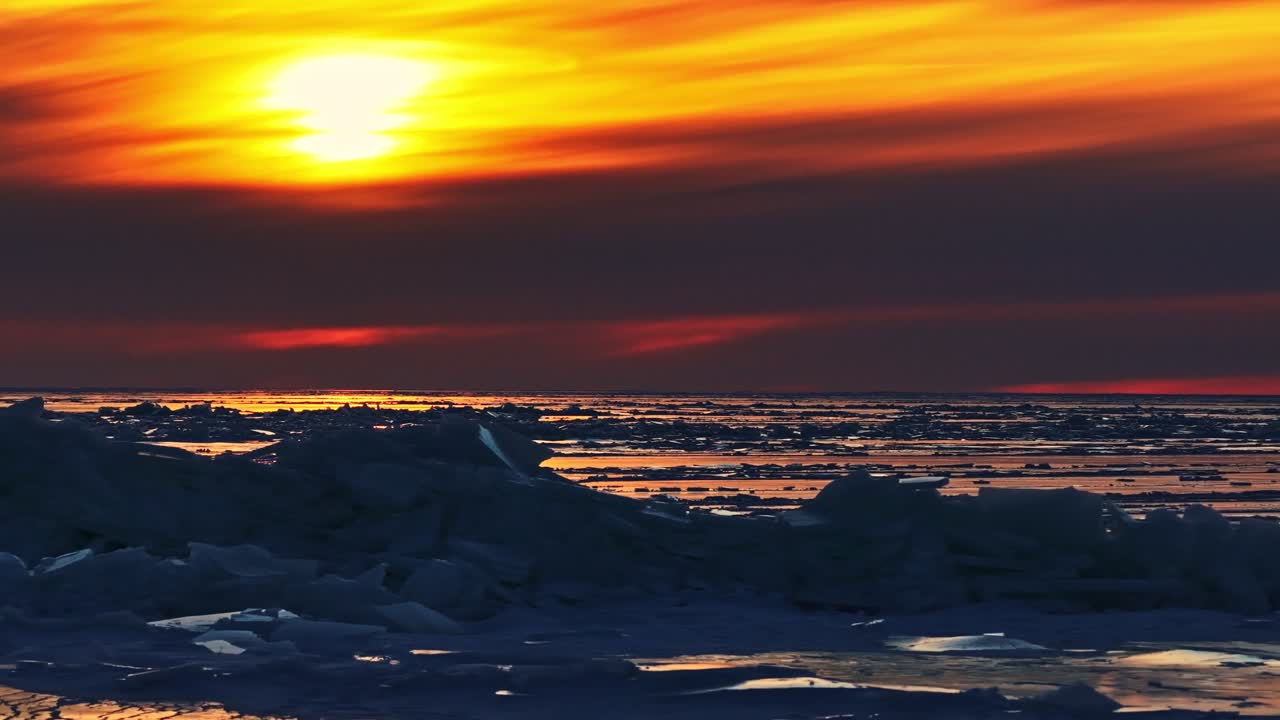 Gefrorenes Meereis spiegelt einen dramatischen Sonnenuntergangshimmel in der Dämmerung wider. Meereisformationen treiben auf dem Wasser und reflektieren goldenes und orangefarbenes Licht der untergehenden Sonne.