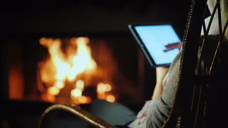 una mujer usa una tableta mientras se sienta en una mecedora junto a la chimenea en casa