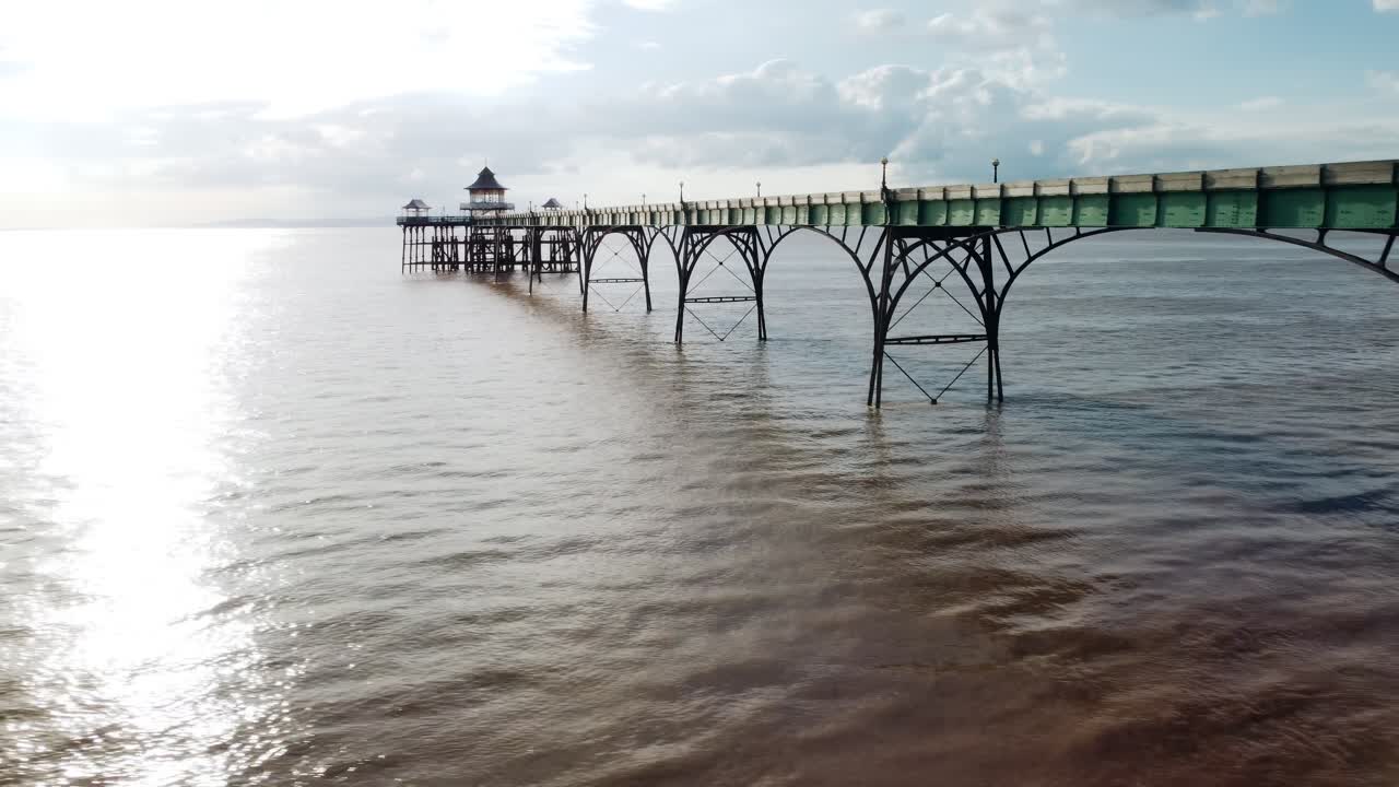 Ripresa con drone che sorvola sotto il Molo di Clevedon al tramonto. Mostra il molo che si estende verso il mare. Il sole si riflette sul mare