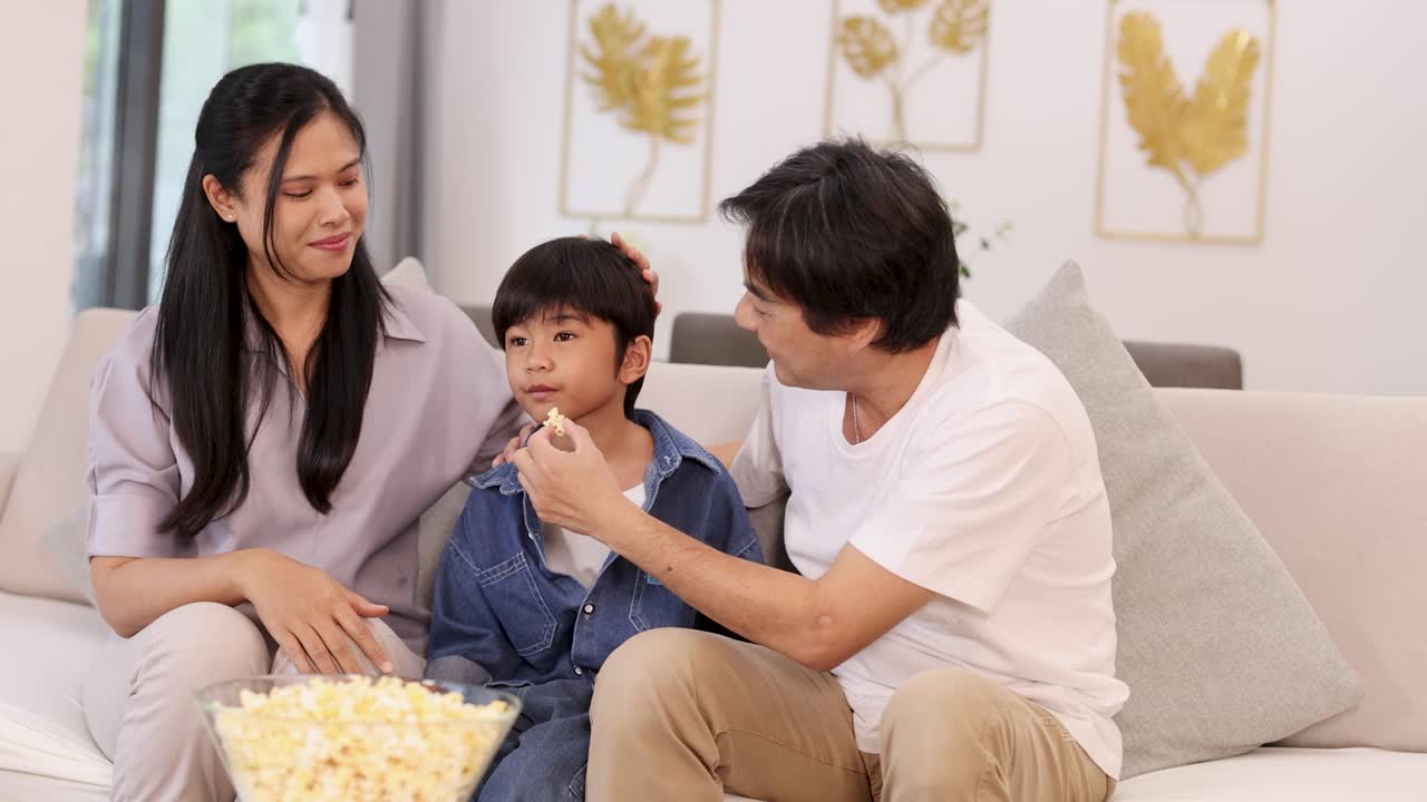Happy Asian family enjoys popcorn and TV together in a cozy, warmly lit living room