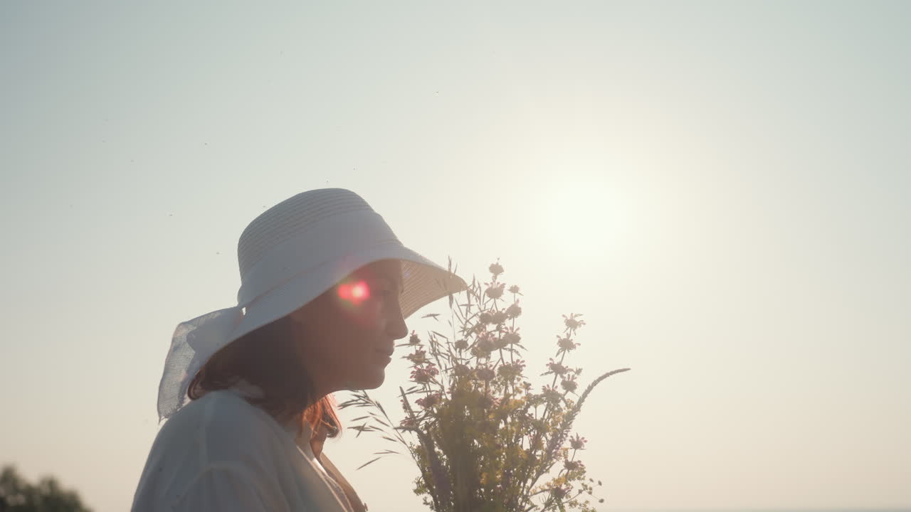 woman in white sun hat stands with wildflowers under bright sunlight, gently inhaling floral scent with relaxed expression as lens flare and morning light create serene countryside