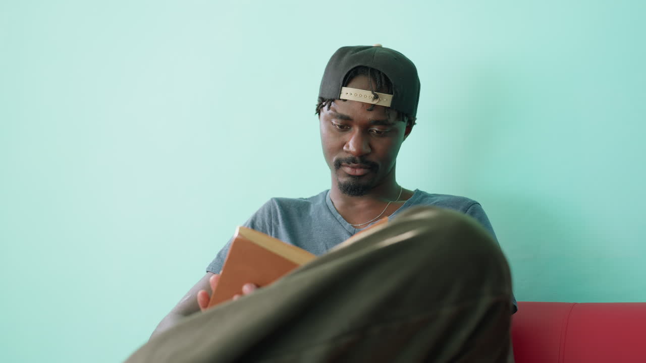 Close up of focused academician in casual shirt and cap sitting on couch with crossed leg, holding open book while reading attentively in bright indoor space with turquoise wall