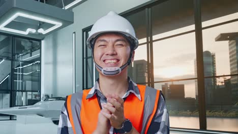 안전 헬을 입은 아시아 남성 엔지니어가 드론 프로젝트에 서서 웃고 손을 박고 있습니다.