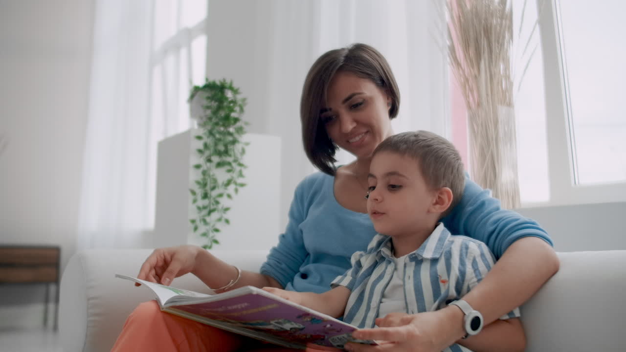 una joven madre con un niño leyendo un libro sentado en un interior blanco brillante de la casa en la sala de estar en el sofá