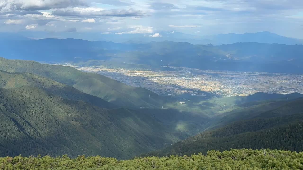 Wide shot overlooking Azumino city from high alpine mountains, with layered green ridges and drifting clouds under a bright sky