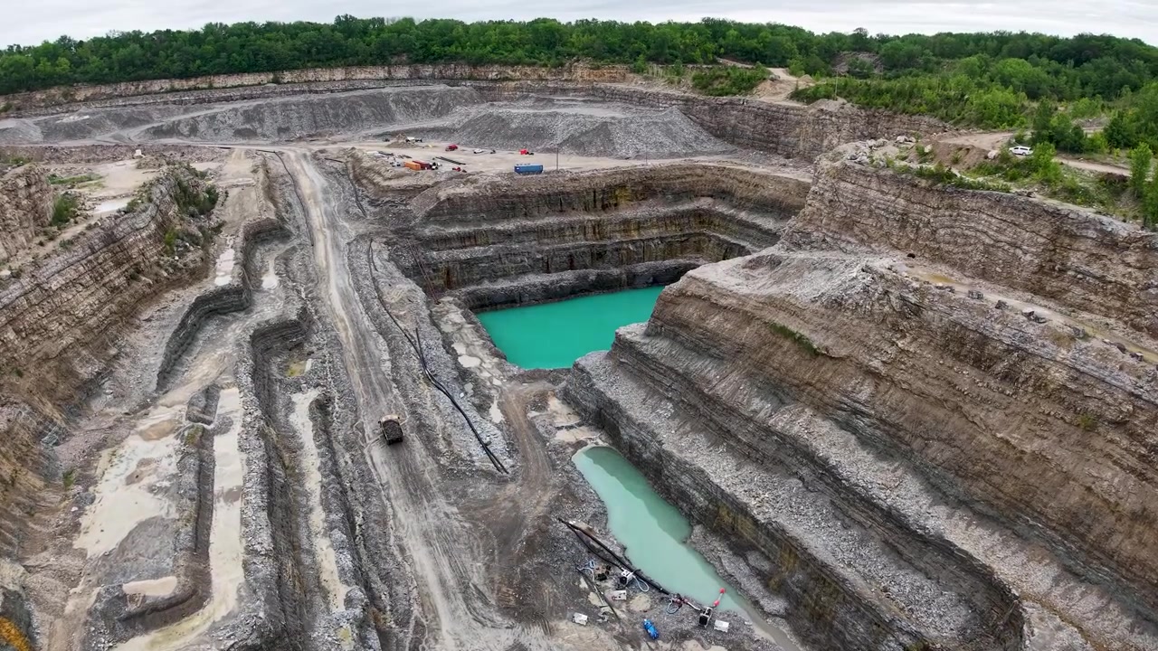 High drone view into a deep terraced quarry with a central haul ramp and turquoise pools