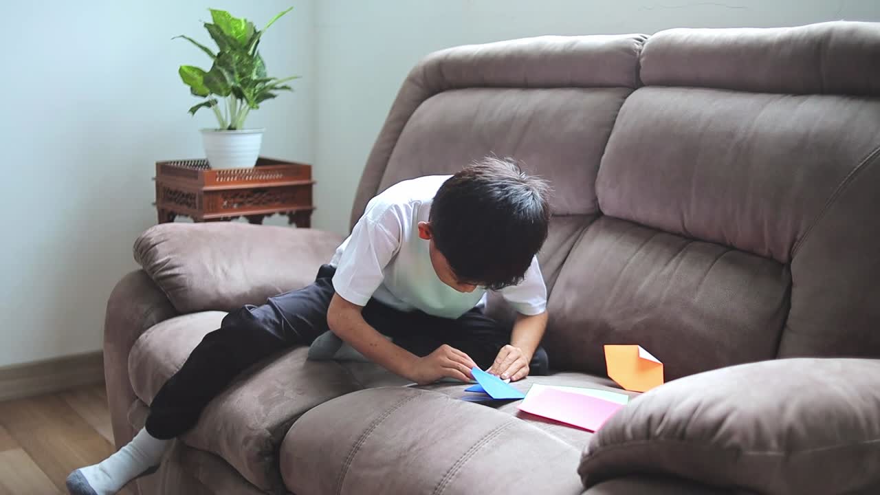 Focused young asian boy creating colorful origami on plush sofa