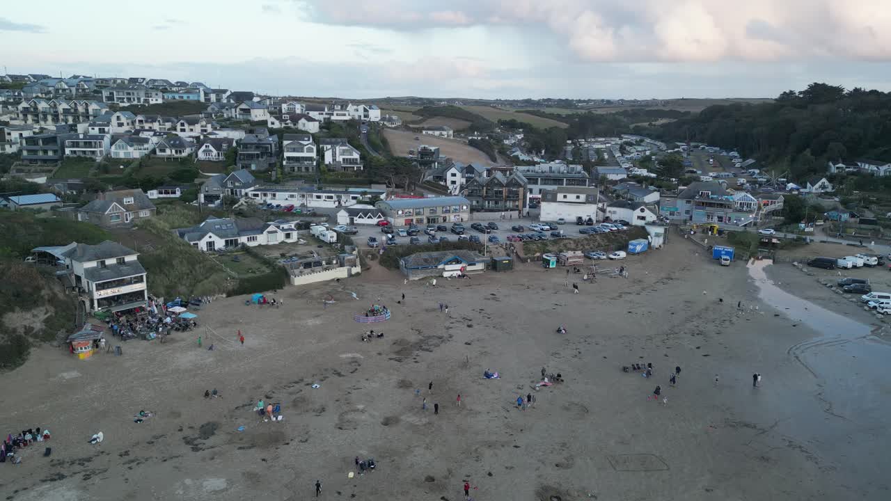 Drone video capturing Polzeath Beach, Cornwall, showcasing a bustling shoreline with people, vibrant homes, and lush hills under soft sunset light, offering dynamic coastal scenery