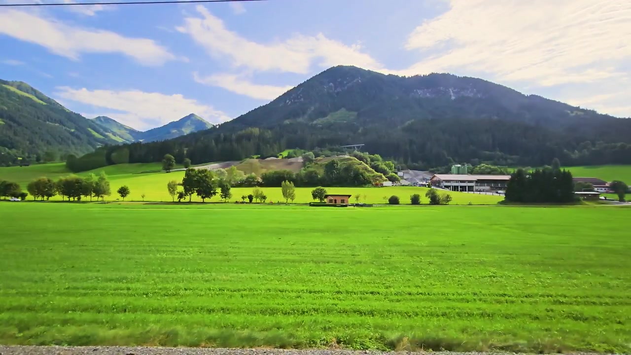 ヨーロッパの明るい夏の雲の下、列車窓から見たオーストリアの田園風景。なだらかな緑の畑、伝統的な農家、森に覆われた丘、そして壮大なアルプスの山々が広がる。