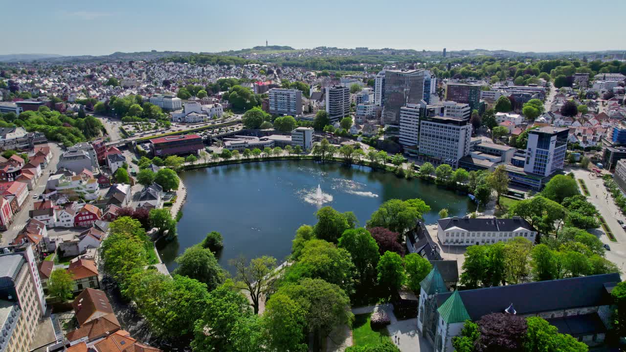 Smooth drone pan around Breiavatnet showcasing the beautiful central fountain and Stavanger’s high-rise buildings in the background on a sunny day.