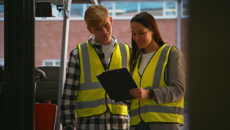 female team leader with clipboard in warehouse with male intern standing by fork lift truck