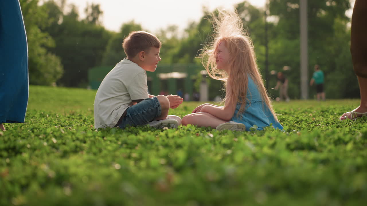 Playmates seated on grass while parents spreading blanket over them in sunny park, face painted girl and boy sharing joyful moment, green clover field background, family outdoor bonding